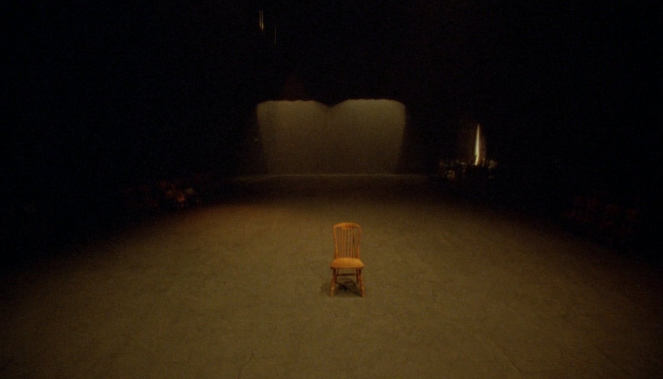 Empty rehearsal room with a single wooden chair center frame, warm amber stage lighting and dark wings receding into shadow